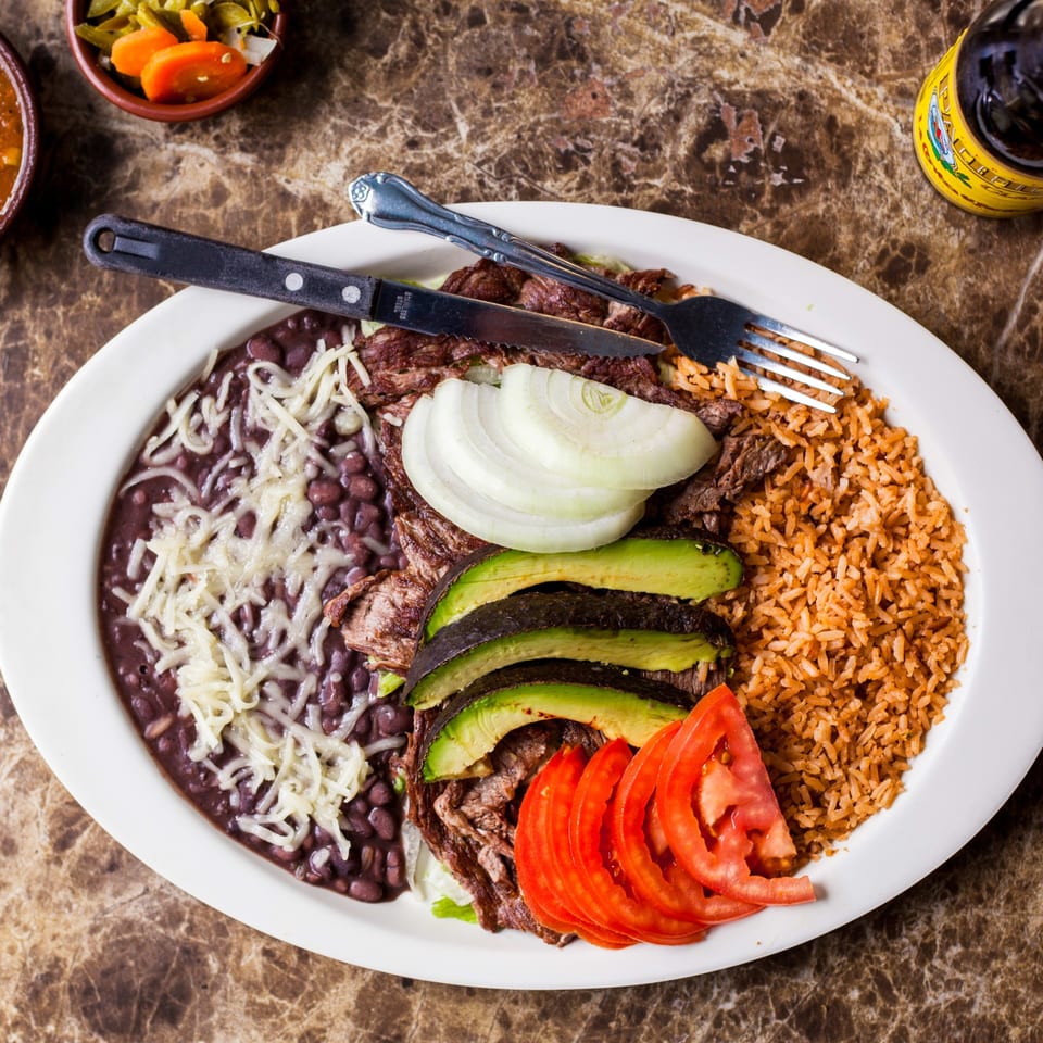 Gorditos' carne asada plate with rice, beans, avocado, tomatoes, and onions on a marble table.